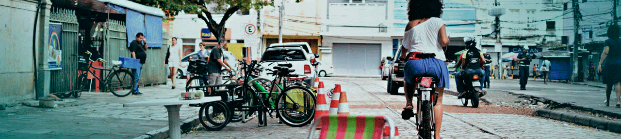 Mulher negra de cabelo crespo volumoso andando de bicicleta com camisa branca de costas no canto direito do banner, passando ao lado de um bicicletário com várias bicicletas e cones que protegem este bicicletário